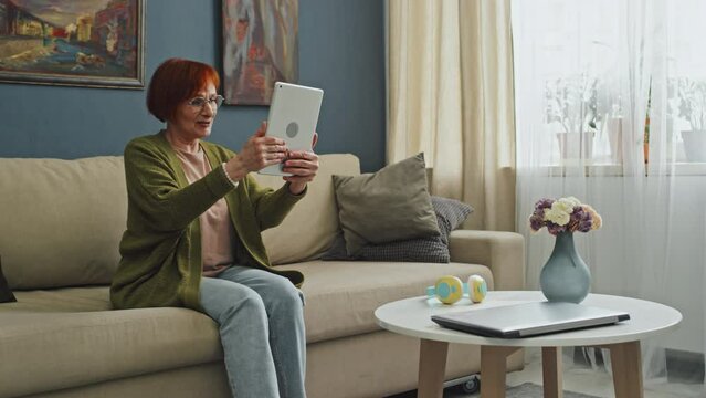 Panning left full shot of cheerful senior woman with red hair sitting on couch in living room holding tablet and having video call with family