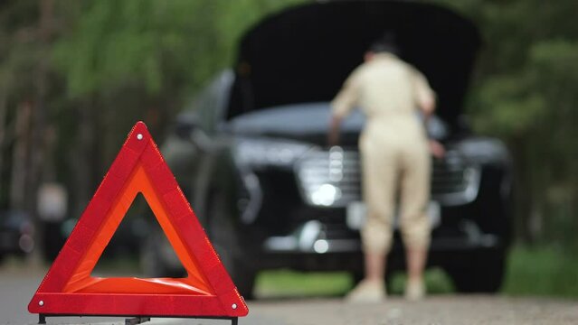 Warning triangle against blurry woman driver trying to fix defect alone in engine of automobile. Businesswoman looking for faults and trying to fix black car
