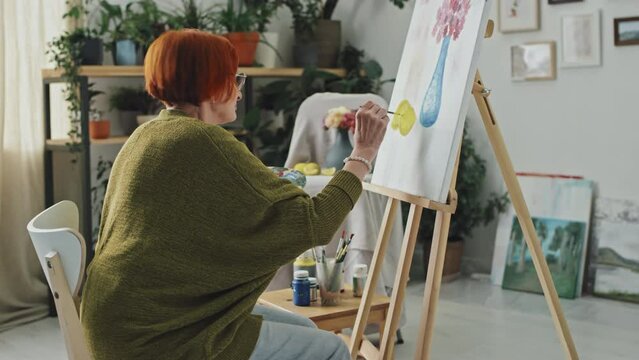 Panning right medium shot of senior woman sitting in front of easel painting colorful still life picture