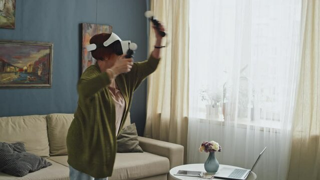 Medium full shot of senior woman wearing VR goggles and holding motion sensors playing video games in middle of living room at daytime