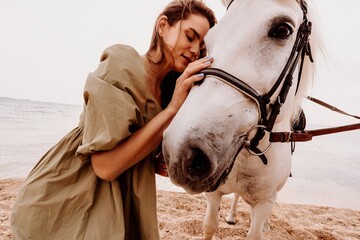 A woman in a dress stands next to a white horse on a beach, with the blue sky and sea in the...