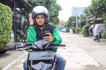 Asian online taxi driver wearing green jacket and helmet feeling bored while waiting order from smartphone