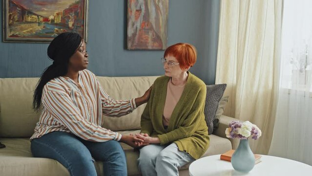 Panning left medium full shot of African American young woman sitting next to and supporting troubled senior lady with red hair in apartment at daytime