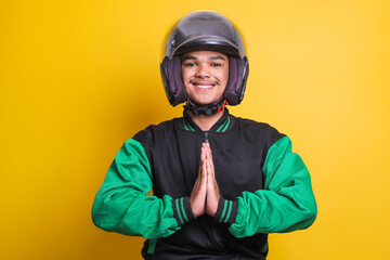 Asian online taxi driver wearing green jacket and helmet smiling and placing hands in greeting or...