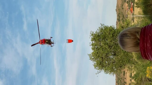 Back view head of unrecognizable blond woman looking at red fire helicopter flying on sky. Vertical video of Caucasian lady watching rescue aircraft outdoors