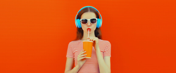 Portrait of cool young woman in headphones listening to music and drinking fresh juice on orange background