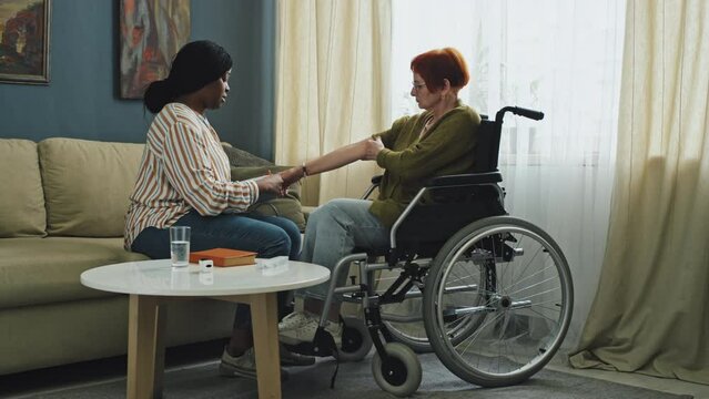 Panning left full shot of senior woman in wheelchair and African American nurse checking her blood pressure in living room at daytime