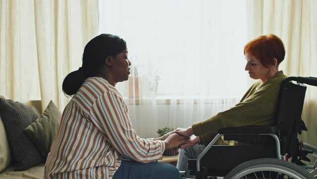 Panning right medium shot of upset senior woman in wheelchair and African American home care assistant calming her down and holding her hand in living room at daytime