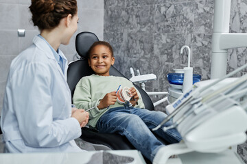 Portrait of smiling little girl sitting in dental chair and listening to female dentist in consultation on tooth health, copy space