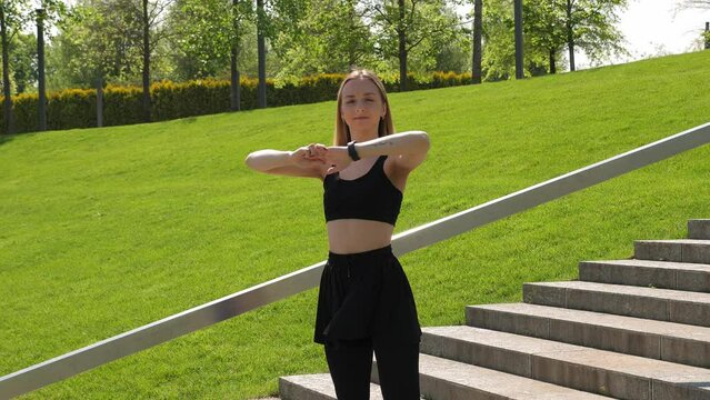 A beautiful young woman is warming up before training in a modern city park on the stairs. Training outdoors in the morning in sunny weather.