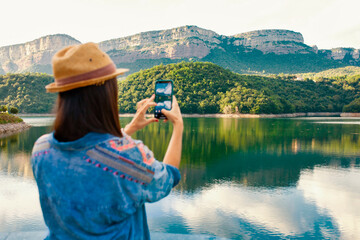 Back view of anonymous young female traveler in stylish clothing and hat taking picture with mobile phone while standing near mountain lake during summer holidays.