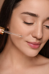 Beautiful woman applying essential oil onto face on brown background, closeup