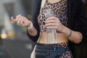 Trendy young woman in leopard clothes drinking water from a reusable glass bottle on a walk. Unrecognizable female person hydrating with mineral water outdoor
