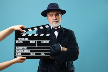 Senior actor performing role while second assistant camera holding clapperboard on light blue...