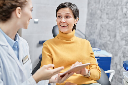 Portrait of smiling mature woman in dental clinic talking to female dentist and holding tablet