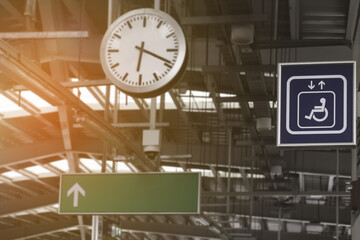 Focus to blue symbol of lift or elevator for disabled person or wheelchair in train station with blurred background of time on clock and white up arrow on empty green direction light box with sunrise.