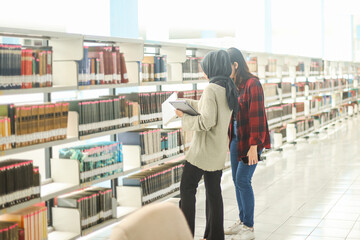 Young Muslim female student with her Asian friend choosing books to read at library for studying, academic research, or school work or project