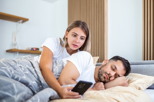 Jealous wife spying the phone of her partner while he is sleeping in a bed at home. Shocked jealous wife spying the phone of her husband while man sleeping in bed at home
