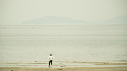 曇り空の広い砂浜を散歩するフレンチブルドッグと飼い主の男性