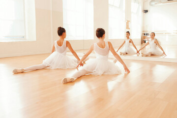 Back view of teen ballerinas sitting in splits on floor and looking in mirror while stretching in dance hall. © carlesmiro