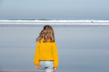 Kid blondie girl standing at the beach looking at the sea in her backs. She is wearing winter clothes, jumper and jeans.