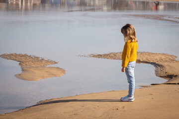 Kid blondie girl standing on the sand beach looking at the sea. She is wearing winter clothes, jumper and jeans.