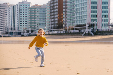 child girl running on the beach with the town of Gijon, Asturias, in the background. She is wearing winter clothes, jumper and jeans. 