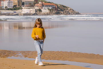 girl walking on the beach with the town in the background. She is wearing winter clothes, jumper and jeans. she is holding a shell in her hands