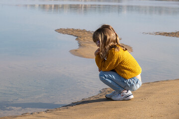 Kid blondie girl sitting on the sand beach looking at the sea. She is wearing winter clothes, jumper and jeans.