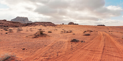 Fototapeta na wymiar Rocky massifs on red orange sand desert, vehicle tracks ground, cloudy sky in background, typical scenery in Wadi Rum, Jordan