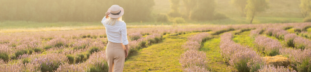 Naklejka na ściany i meble Beautiful young healthy woman with a white dress running joyfully through a lavender field holding a straw hat under the rays of the setting sun