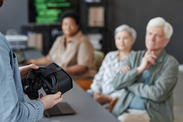 Closeup of man holding VR headset presenting virtual reality technology to group of senior people,...