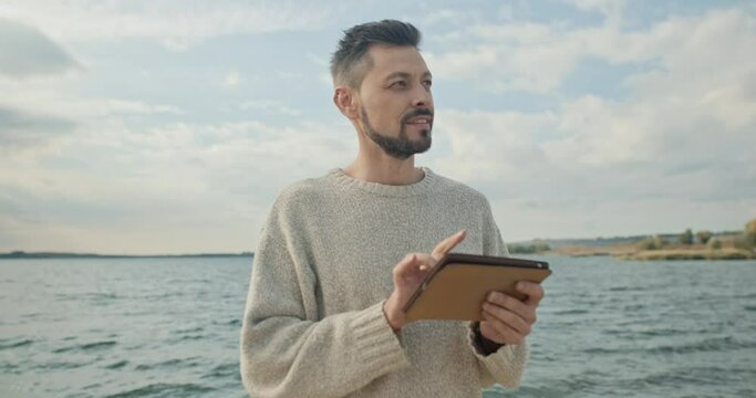 Man stands at background of lake and sky. Male smiles and looks at tablet. Guy holding gadget and texting. Man going out into nature and communicates with someone by device.