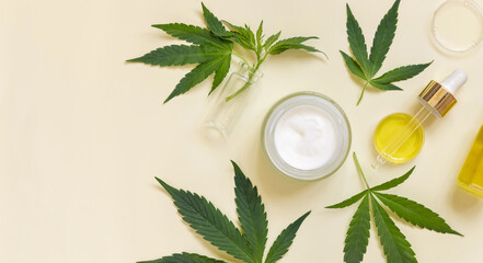 Pipette and opened cream jar near green cannabis leaves top view on beige, CBD cosmetics