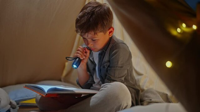 Child boy reading book with fairy tales or scary bed stories with flashlight inside play tent wigwam