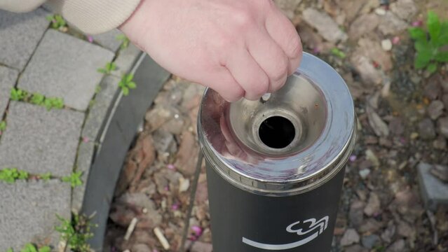 Smoker hand throws cigarette butt into street ashtray in designated outdoor smoking area.