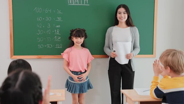 Student girl answering at blackboard to woman teacher of math in the classroom. Cheerful diverse student clap hands while friend writing correct answer on blackboard at school