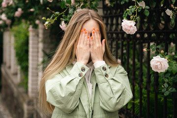 Young blonde woman with long hair near the fence with a climbing rose covers her face with her hands with orange manicure, mental health concept, outdoor portrait.