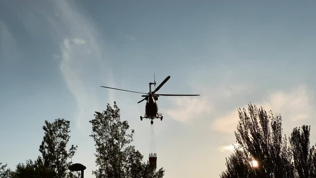 Silhouette of fire helicopter flying down to water reservoir in sunset sunbeam. Back view aircraft chopper collecting water for fire extinguish outdoors at sundown
