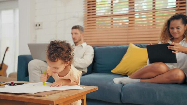 Modern parents working by computer and tablet while cute little daughter drawing a picture in living room at home. Happy father mother and daughter to spend time together at home. Family concept