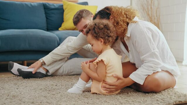 Parents warmth by embracing and teasing little daughter while spend time together on floor in living room at home. Mom and Dad using spare time with baby together at warmth house. Family concept.