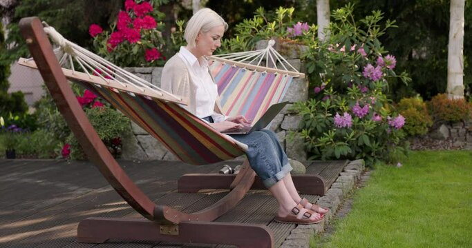 Woman sitting on hammock typing on laptop in backyard terrace