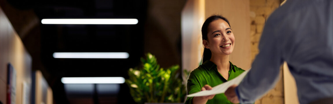 Young cheerful asian woman giving some documents to her colleague and smiling while standing together in the modern office