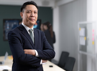 Portrait of Asian businessman standing in conference room arms crossed. Confident smart entrepreneur wears suit looking at camera smiling. Handsome businessperson meeting with colleagues in workplace.
