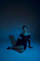 Fototapeta na wymiar full length of appealing asian woman in black cocktail dress, fishnet tights and long gloves looking away while sitting on blue background with cyan lighting effect, fashion model