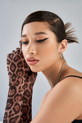 portrait of beautiful and dreamy asian woman in animal print gloves and silver accessories, with trendy hairstyle and bold makeup on grey background, stylish spring, fashion shoot