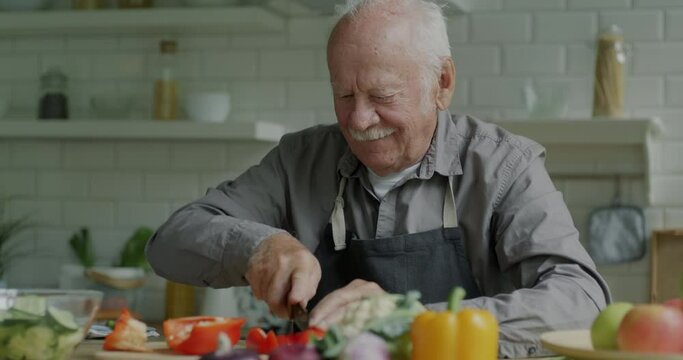 Portrait of cheerful elderly man cutting vegetables making salad indoors in kitchen at home. Dish recipe and food preparation concept.