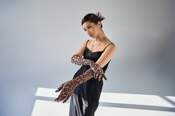 youthful asian fashion model with trendy hairstyle and bold makeup posing in trendy spring outfit, black strap dress and animal print gloves on grey background with lighting