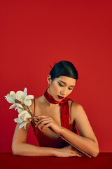 Fototapeta premium youthful fashion, trendy spring concept, charming asian woman in neckerchief and strap dress sitting at table with white blooming orchid on red background