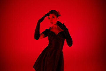 young asian woman in black long gloves and elegant cocktail dress standing in expressive pose and looking away on vibrant background with red lighting effect, youth culture, fashionable spring